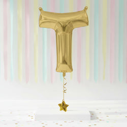 Gold foil balloon shaped like a letter 'T' with a star on a pastel striped background